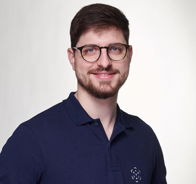 Professioneller Mann mit Brille und dunklem Polo vor hellem Hintergrund.