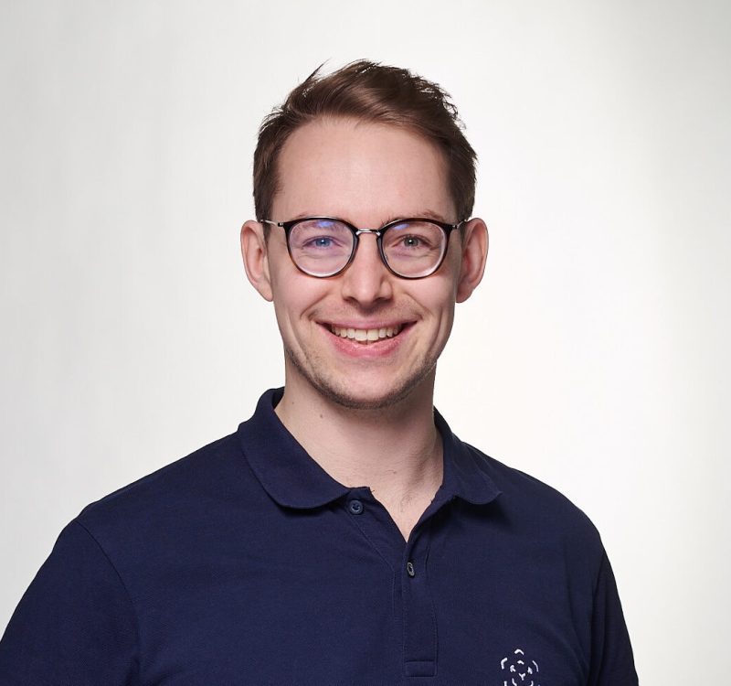 Junger Mann mit Brille und dunklem Polo vor hellem Hintergrund.